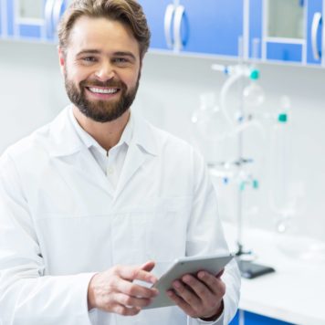 Positive mood. Delighted handsome nice biologist smiling and looking at you while holding a tab let in his hand
